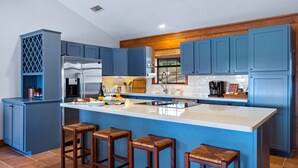 A modern kitchen with blue cabinets, a stainless steel refrigerator, a large island with stools, and a window above the sink. - Lodge II | Lake Views | Hot Tub | Pool + River Access (Ingram)