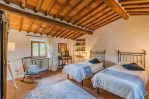 Quarto aconchegante com duas camas, vigas de madeira no teto, uma cadeira de balanço e uma escrivaninha com abajur.
