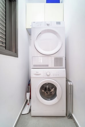 Un appareil combiné lave-linge et sèche-linge à chargement frontal, de couleur blanche, doté d’un panneau de commande, installé dans un coin d’une pièce avec une fenêtre et un radiateur.