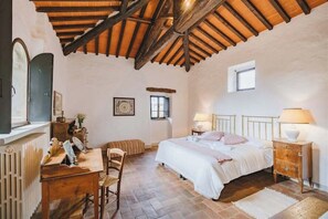 Un dormitorio con techo de madera, una cama con ropa blanca, una mesita de noche con lámpara, un aparador de madera y una ventana.