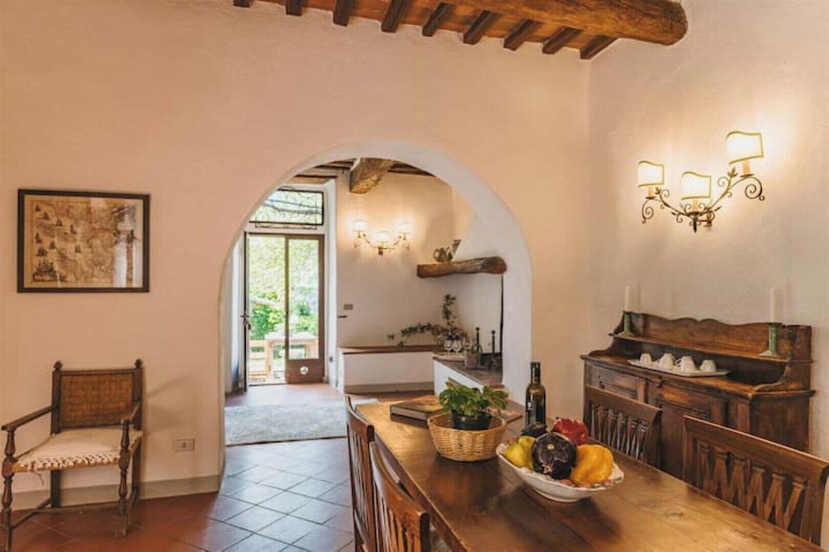 A dining area with a wooden table, chairs, and a fruit bowl. An archway leads to a kitchen with a sink and a hanging light fixture.