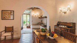 Un coin repas avec une table en bois, des chaises et un panier de fruits. Un arc donne accès à une cuisine équipée d’un évier et d’un luminaire suspendu.