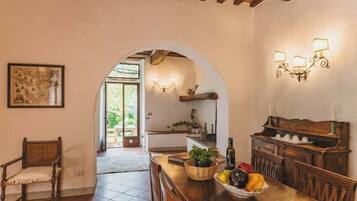 A dining area with a wooden table, chairs, and a fruit bowl. An archway leads to a kitchen with a sink and a hanging light fixture.