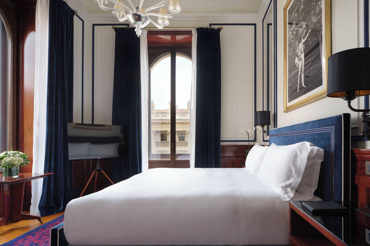 A hotel room with a large bed, a desk, a chair, a lamp, and a view of a building through a window.