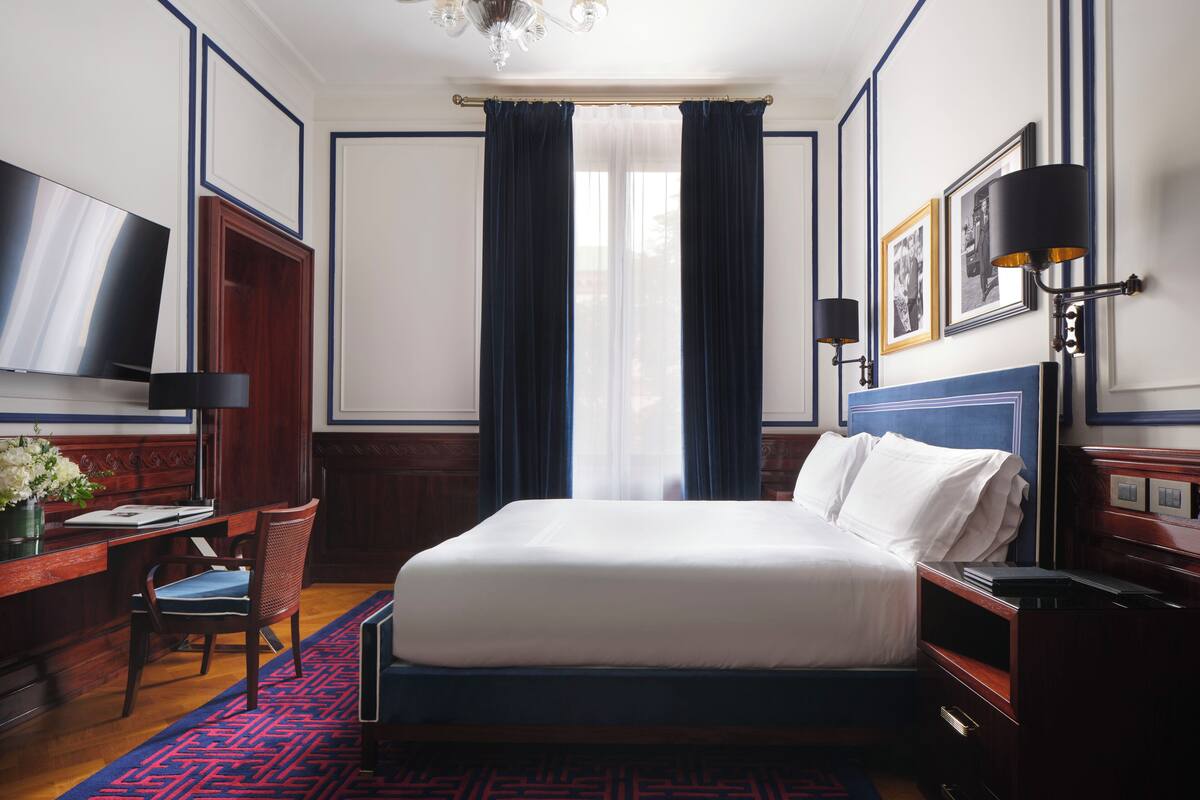 A hotel room with a large bed, a desk with a chair, a television, a chandelier, and a wall-mounted lamp.