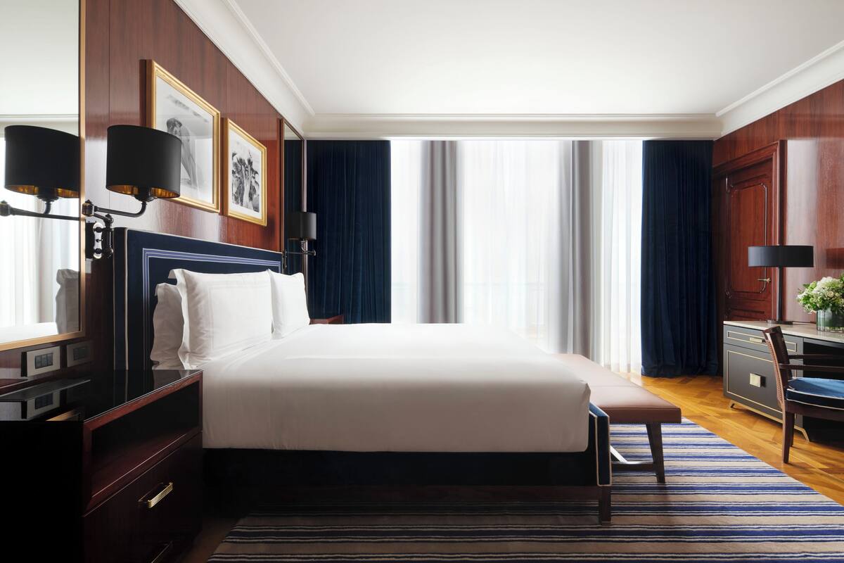 A hotel room with a large bed, a desk with a lamp, a chair, and a window with curtains.