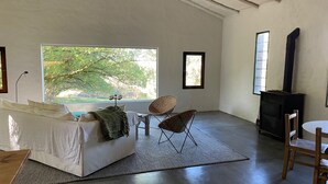 A spacious room with a large window, a white sofa, two wicker chairs, a small table, and a black stove. - Minutos a Jose Ignacio, La Casita De Infinita, en el campo, cerca la playa! (Faro de José Ignacio)