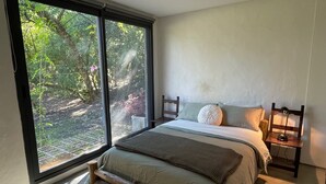 A bedroom with a large window, a bed with a wooden headboard, a bedside table with a lamp, and a view of greenery outside. - Minutos a Jose Ignacio, La Casita De Infinita, en el campo, cerca la playa! (Faro de José Ignacio)