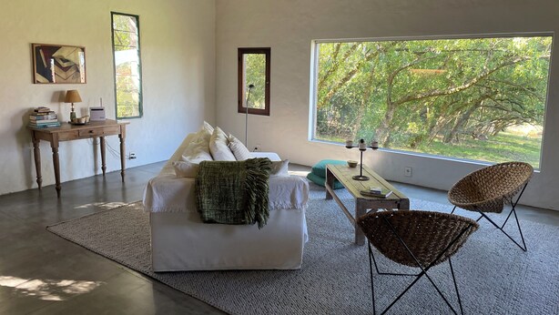 A modern living room with a sofa, a coffee table, a chair, and a desk with a lamp and books. There is a large window with a view of trees. - Minutos a Jose Ignacio, La Casita De Infinita, en el campo, cerca la playa! (Faro de José Ignacio)
