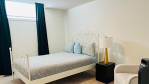 Une chambre à coucher avec un lit en métal blanc, une table de chevet avec une lampe, une chaise et une fenêtre avec des rideaux.