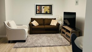 Un salon avec un canapé marron, un fauteuil blanc, un téléviseur et un tableau abstrait encadré accroché au mur.