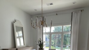 Un comedor con mesa de madera, sillas blancas tapizadas, una araña, un ventanal con cortinas y vistas al exterior.