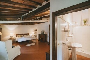Quarto com cama, mesas de cabeceira e um criado-mudo. Há um banheiro com pia e espelho. O quarto possui vigas de madeira no teto.