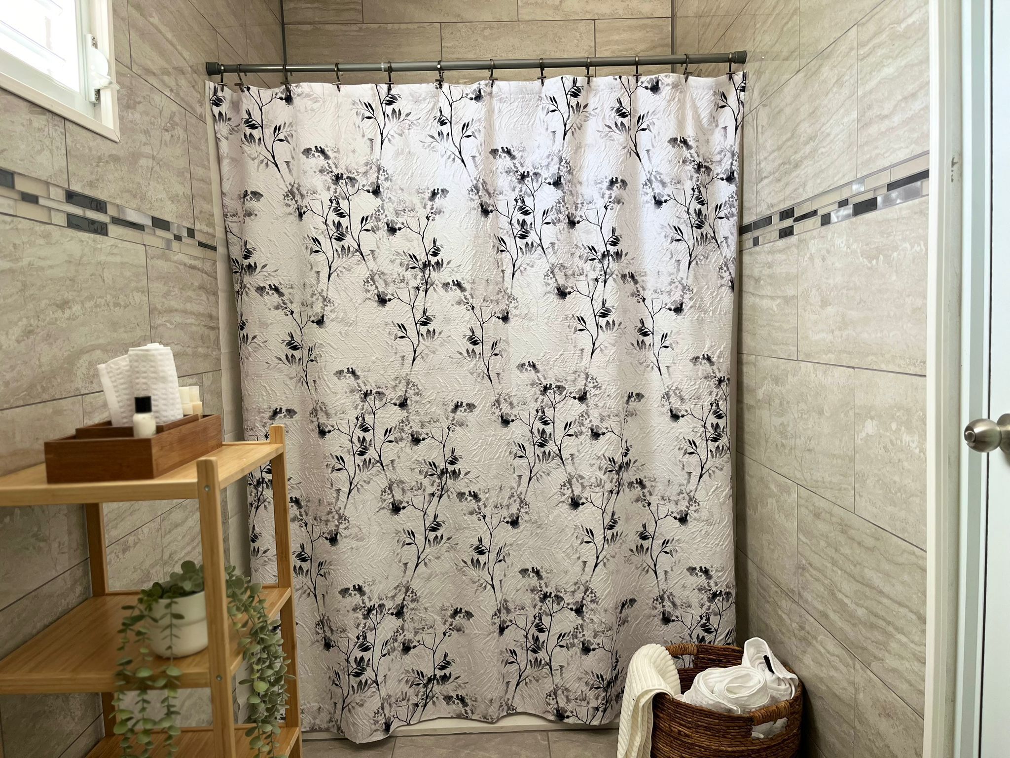 Une salle de bain avec un rideau de douche orné d’un motif floral, une étagère en bois contenant des articles de toilette et un panier en osier rempli de serviettes.