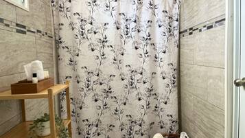 Une salle de bain avec un rideau de douche orné d’un motif floral, une étagère en bois contenant des articles de toilette et un panier en osier rempli de serviettes.