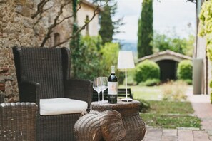 Una mesa de mimbre con una botella de vino y copas, una lámpara blanca y una silla de mimbre con cojín.