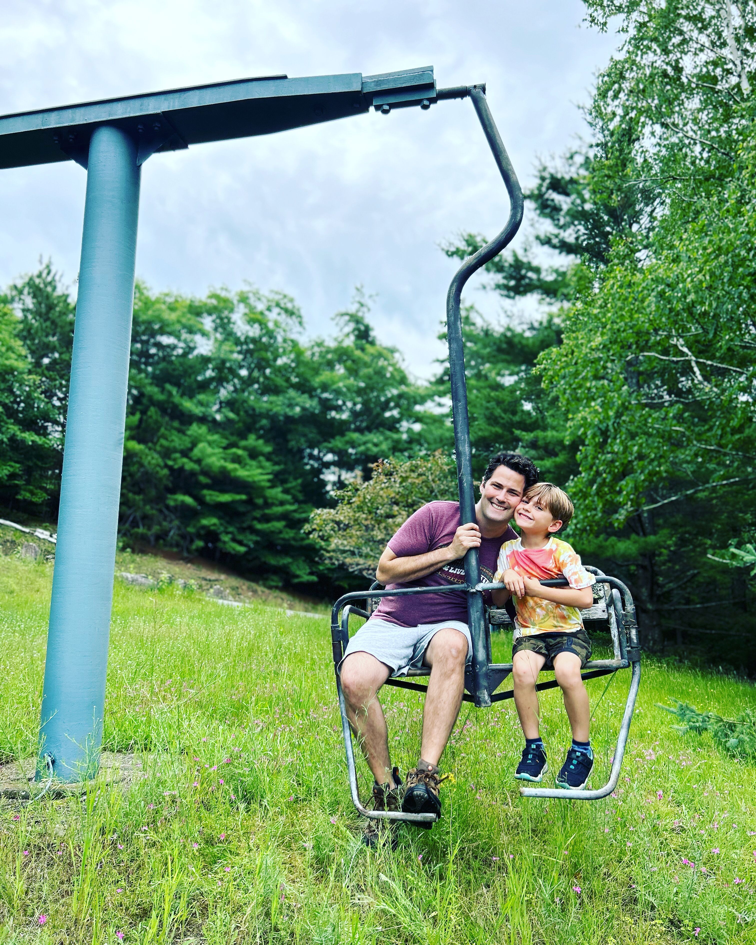 A man and a child on a swing set in a grassy area with trees in the background.