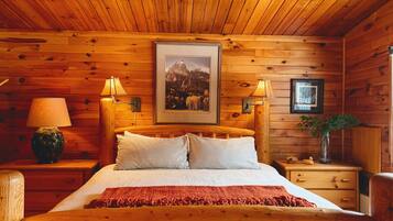 Una habitación de cabaña de madera con una cama, dos lámparas de noche, un cuadro enmarcado en la pared, una mesita con un florero y una mesilla de noche.