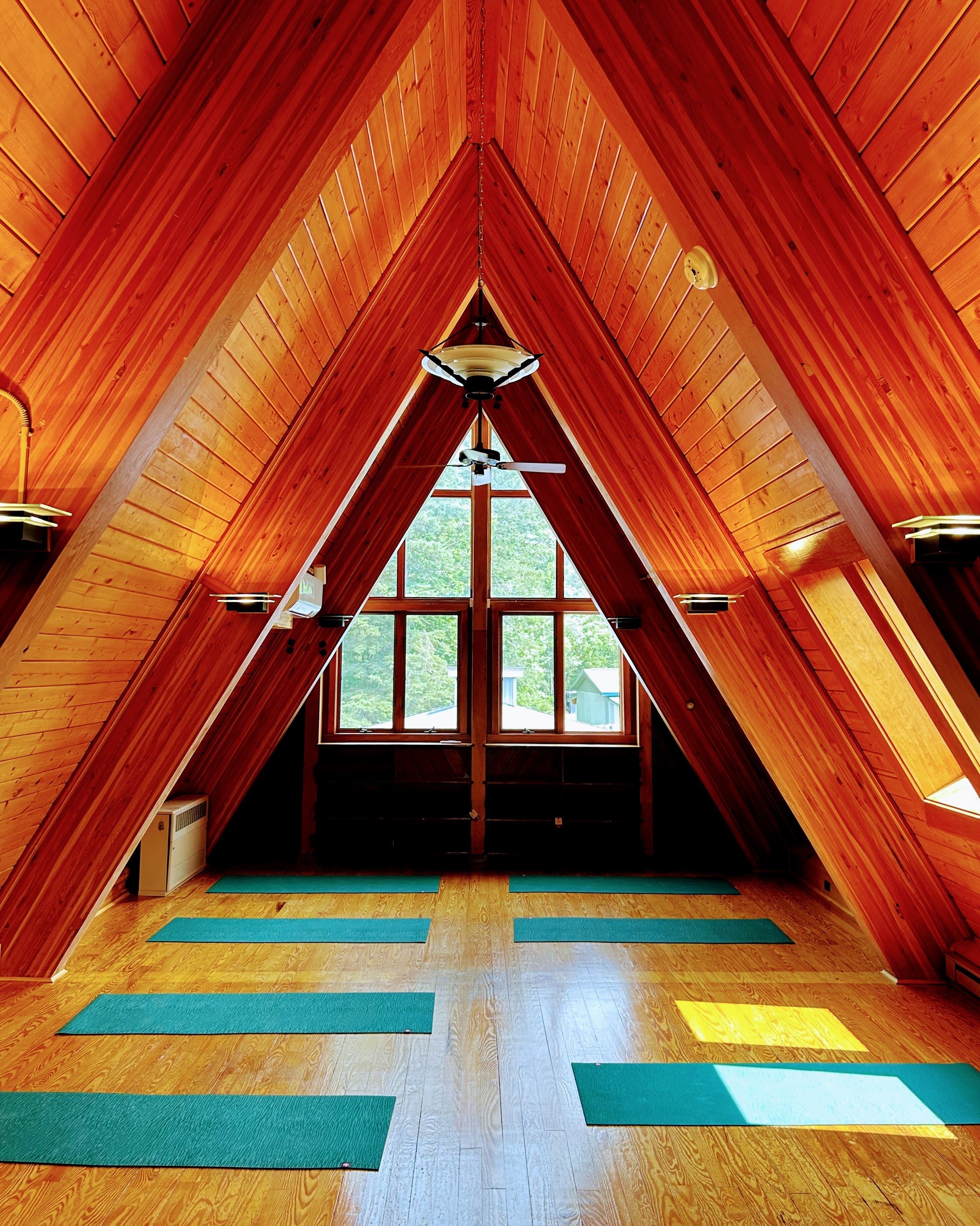 A high-ceilinged attic room with wooden walls and a sloped roof, featuring large windows and a central light fixture.