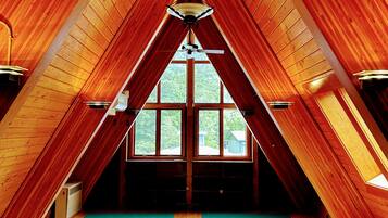 A high-ceilinged attic room with wooden walls and a sloped roof, featuring large windows and a central light fixture.