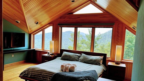 A bedroom with a sloped ceiling, large windows, a bed with pillows, a bedside table, and a lamp. - NewVida Preserve (Jay)