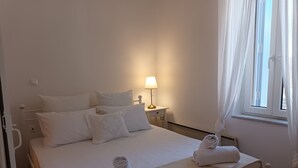 A neatly made bed with white linens and pillows, a bedside lamp, and a window with curtains. - A fully air con 2-bedroom apartment,a cosy living room,fully equipped kitchen. (Tinos)