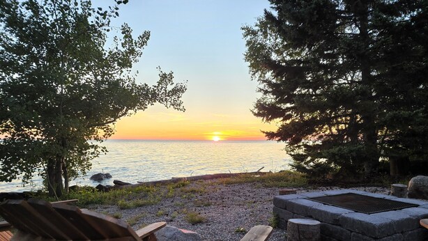 A wooden chair and a stone fire pit by a pebbled beach with a sunset view.