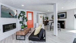 Un salon moderne avec un canapé en cuir noir, une table basse en bois et une grande plante en pot. Une porte rouge et un miroir à cadre blanc sont visibles à l’arrière-plan.