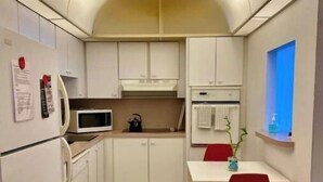 Cocina compacta con armarios blancos, microondas, tetera y comedor con una silla y mesa rojos.
