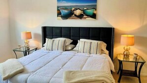 Un cuarto de hotel con una cama grande, dos mesitas de noche con lámparas, un cuadro de barcos en la pared y vista al mar.