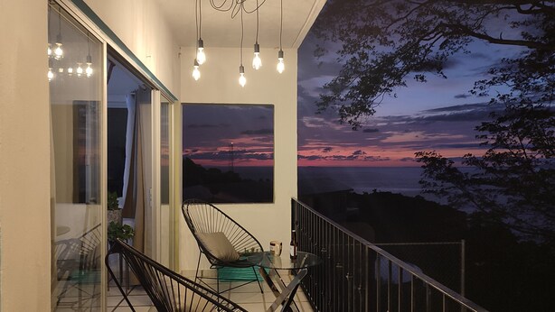 A balcony with a glass door, a black chair, a glass table, and a view of a sunset over the ocean. - Mismaloya Beach (Puerto Vallarta)