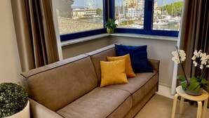 A living room with a brown sofa, two cushions, a small plant, and a view of a marina with boats and a Ferris wheel. - Beautiful flat on the second floor with lift with beautiful modern fittings and large windows with a splendid view of the Viareggio dock just a few minutes' walk from the promenadeand beach.Elegantly furnished it consists of a living room with modern wal (Viareggio)