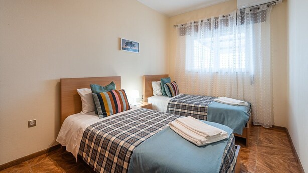 Ein Hotelzimmer mit zwei Betten, einem kleinen Nachttisch mit Lampe, einem Fenster mit durchsichtigen Vorhängen und einer Klimaanlage an der Wand.