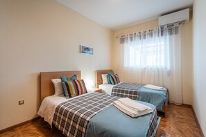 A hotel room with two beds, a small bedside table with a lamp, a window with sheer curtains, and an air conditioning unit on the wall.