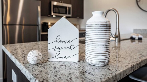 A kitchen counter with a decorative house-shaped sign, a textured vase, and a marble-like object.
