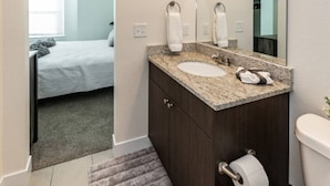 A bathroom with a sink, mirror, toilet, and a bed visible in the adjacent room.