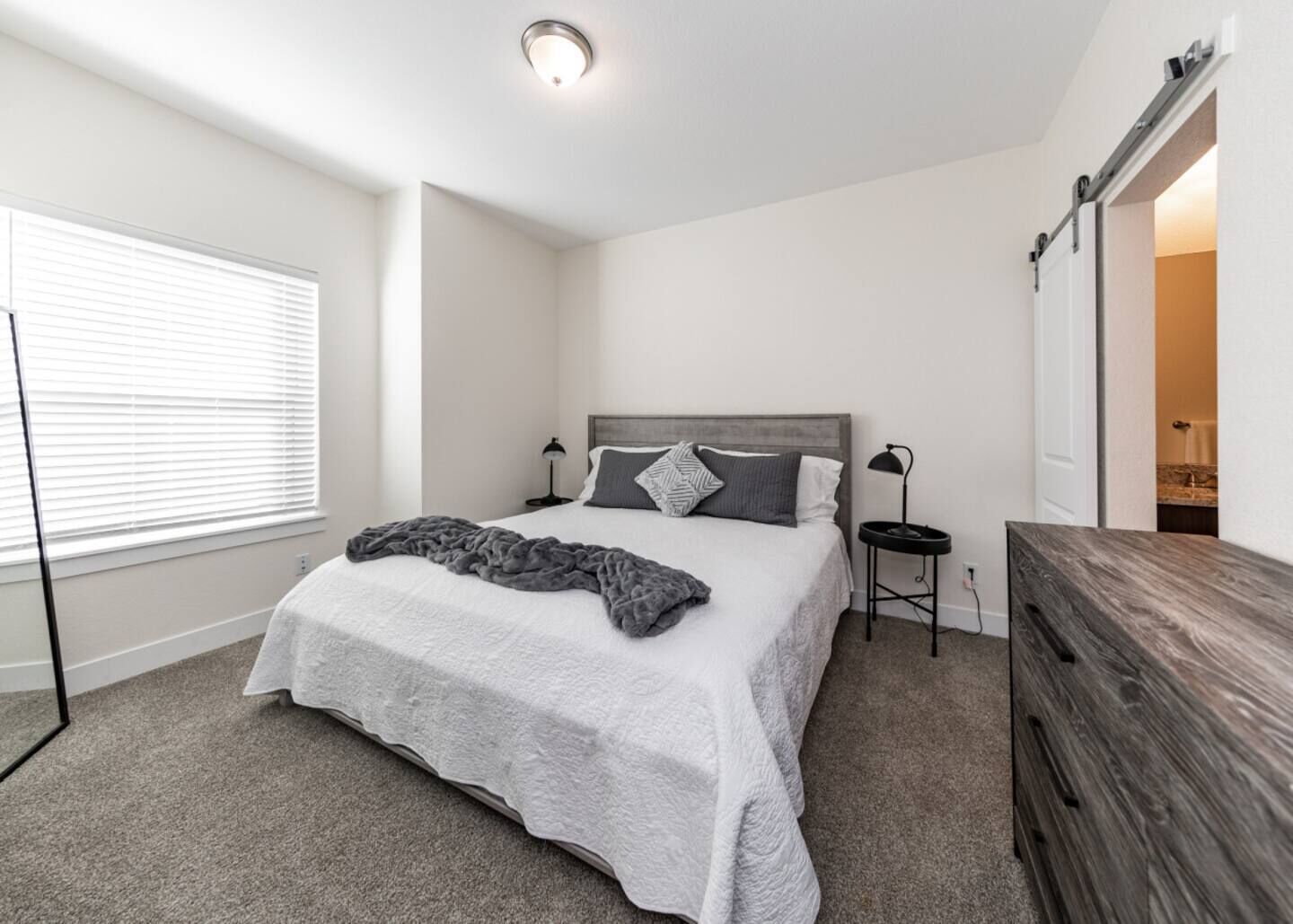 A bedroom with a bed, bedside tables, a dresser, a window with blinds, and a door leading to another room.