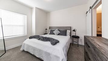A bedroom with a bed, bedside tables, a dresser, a window with blinds, and a door leading to another room.