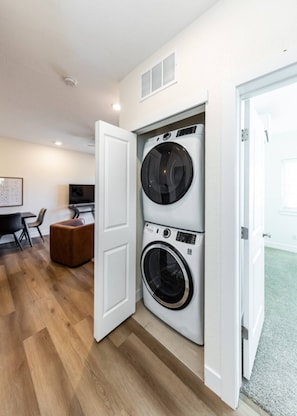 A laundry room with two stacked washing machines, a wooden floor, a white door, and a small seating area with a chair and a table.