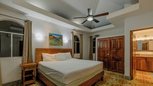 A bedroom with a wooden bed, a nightstand, a ceiling fan, and a bathroom with a sink and mirror. - Casa de Amor B-58                                                             . (Villa El Carmen)