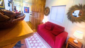 A living room with a red sofa, a wooden coffee table, a patterned rug, a wooden partition, a wall clock, a mirror, and a window with blinds. - The Zanzibar Beach House - North (Dongwe)