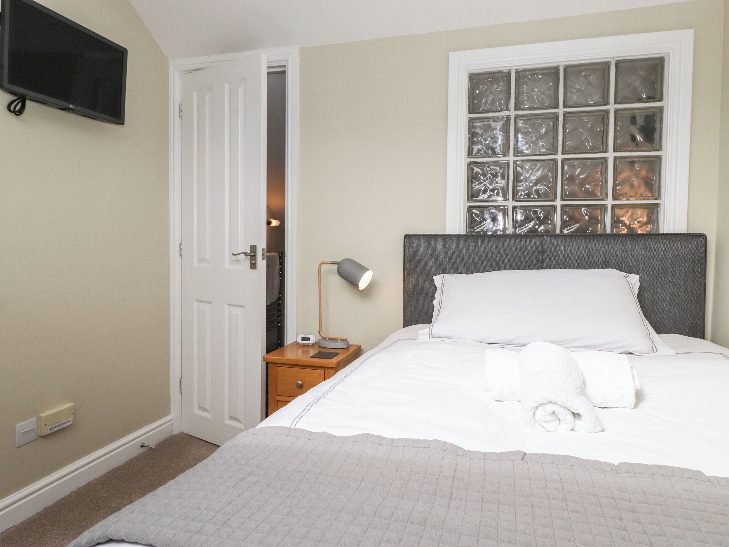 Een net opgemaakt bed met witte lakens, een bedlampje en een raam met decoratieve glaspanelen.