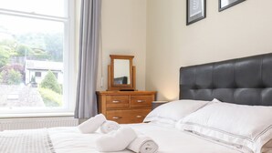 A neatly made bed with white linens and rolled towels, a wooden dresser with a mirror, and a view of greenery outside the window.