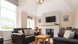 A living room with two dark leather sofas, a wooden coffee table, a fireplace, a television mounted on the wall, and a bookshelf.