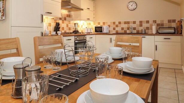 A modern kitchen with white cabinets, a wooden dining table set with white plates, bowls, and glasses, and a tiled backsplash.