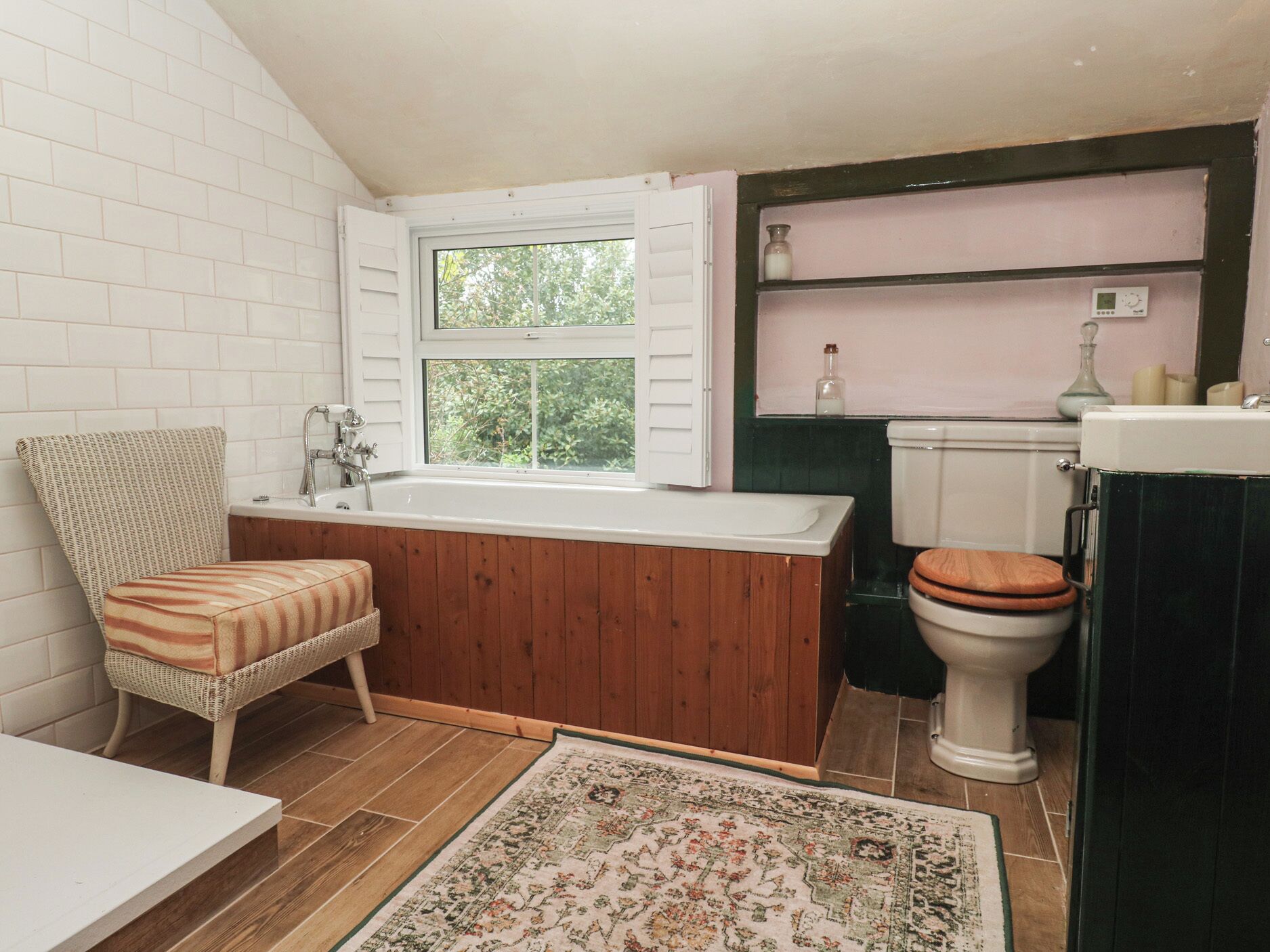 A bathroom with a wooden vanity, a white bathtub, a wicker chair, a toilet with a wooden seat, and a window with shutters.