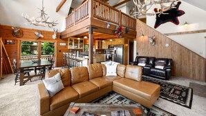 Un vaste espace de vie comprenant un grand canapé d’angle, une table à manger en bois avec des chaises, une cuisine en arrière-plan et un balcon doté d’une balustrade en bois.