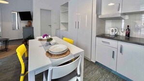 Una cuina moderna amb armaris blancs, una taula de dinar amb cadires grogues i un televisor penjat a la paret.