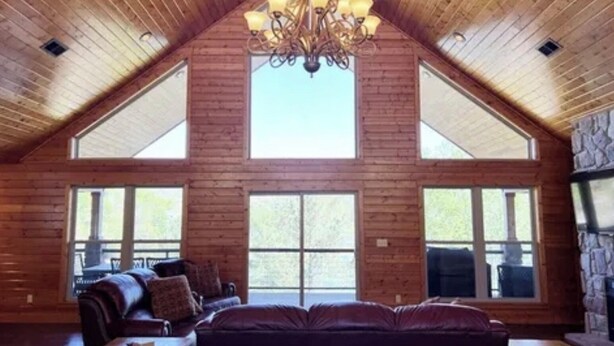 A spacious living room with a high ceiling, wooden walls, and a large chandelier. There are multiple windows providing natural light and a stone fireplace. - SPACIOUS Lakeside Home in the perfect LOCATION near all the action! (LaFollette)