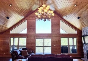 A spacious living room with a high ceiling, wooden walls, and a large chandelier. There are multiple windows providing natural light and a stone fireplace. - SPACIOUS Lakeside Home in the perfect LOCATION near all the action! (LaFollette)